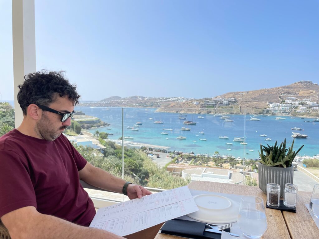 A person sitting at a table with a view of the water and boats in the background Description automatically generated